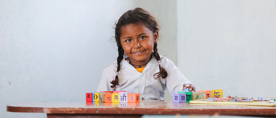 Ei smilende, nepalsk skolejente sitter ved pulten sin og lager ord med bokstavklosser. 